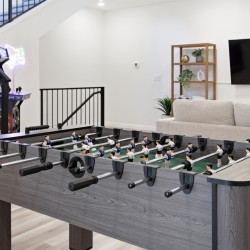 Foosball table in a modern basement recreation room with stairs, arcade machine, and a beige sofa with a wall-mounted TV in the background