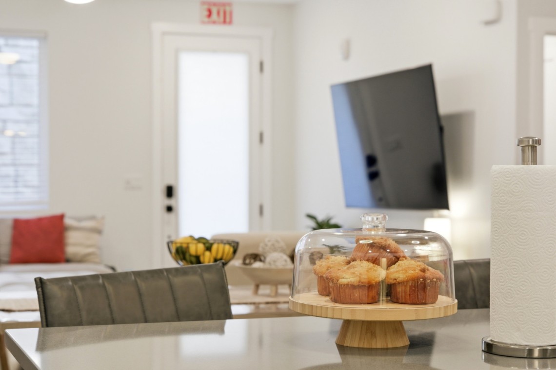 Muffins under a glass dome on a wooden cake stand atop a kitchen counter, with a paper towel roll nearby.