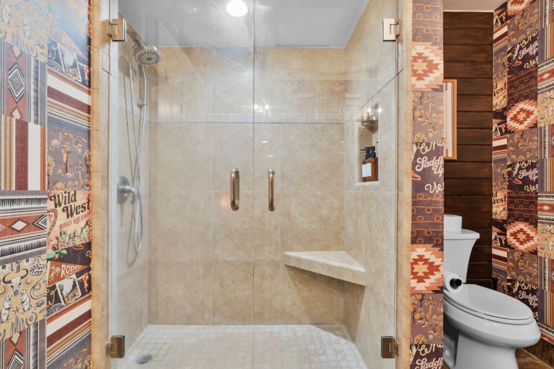 Glass-front walk-in shower with beige tile, chrome fixtures, and a built-in bench; Wild West patterned wallpaper surrounds a toilet on the right.