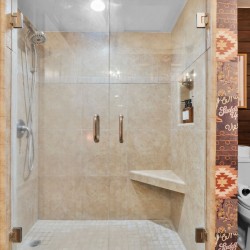 Glass-front walk-in shower with beige tile, chrome fixtures, and a built-in bench; Wild West patterned wallpaper surrounds a toilet on the right.