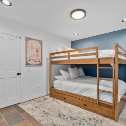 Wooden bunk bed with white bedding against a blue accent wall in a bright kids' room, with a potted plant and rug nearby.