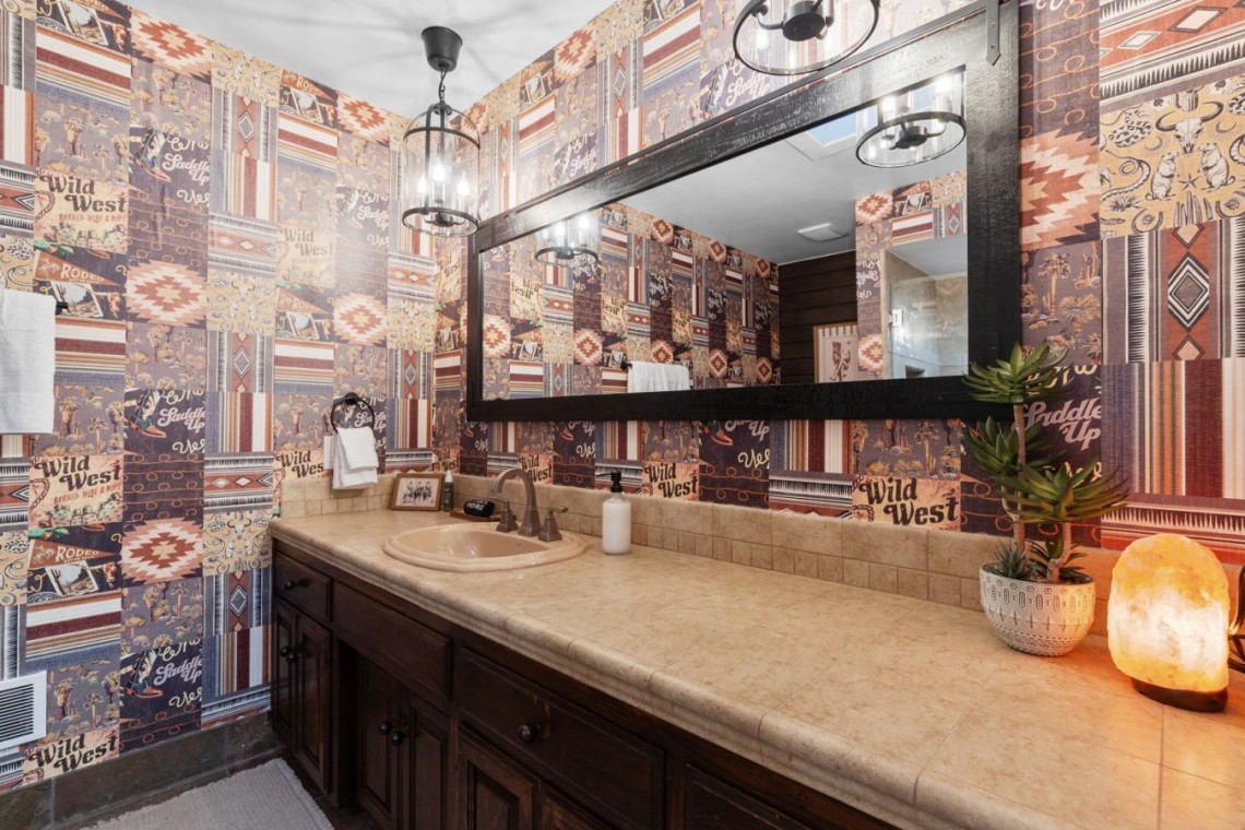 Bathroom with a dark wooden vanity, beige countertop and oval sink, a large black-framed mirror, Western patchwork wallpaper, white towels, a plant, and a glowing Himalayan salt lamp on the counter.