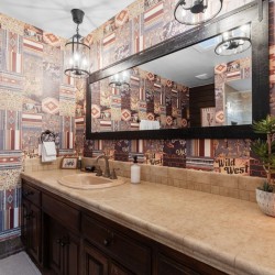Bathroom with a dark wooden vanity, beige countertop and oval sink, a large black-framed mirror, Western patchwork wallpaper, white towels, a plant, and a glowing Himalayan salt lamp on the counter.