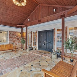 Covered brick entryway with dark double doors, wooden beams, and potted plants, welcoming visitors to a home.
