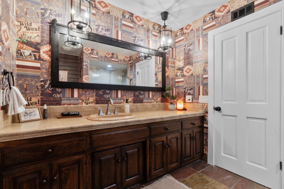 Bathroom with dark wood vanity, beige countertop, and a large black-framed mirror reflecting a doorway and back wall. Wallpaper features patchwork western patterns in warm tones; towel, framed photo, and small plant on counter add decor.