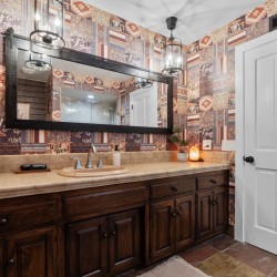 Bathroom with dark wood vanity, beige countertop, and a large black-framed mirror reflecting a doorway and back wall. Wallpaper features patchwork western patterns in warm tones; towel, framed photo, and small plant on counter add decor.