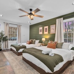 Two twin beds with olive-green throws and orange pillows in a bright bedroom with a green accent wall and framed art above.