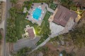 Aerial view of a large estate with a rectangular blue pool, brown-roofed house, green lawns, and a curved driveway surrounded by trees .