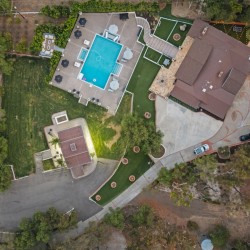 Aerial view of a large estate with a rectangular blue pool, brown-roofed house, green lawns, and a curved driveway surrounded by trees .