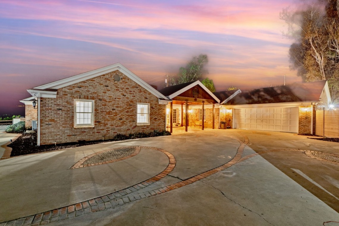 Brick single-story home with a two-car garage at sunset; faint smoke rising from the roof and warm porch lights.