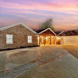 Brick single-story home with a two-car garage at sunset; faint smoke rising from the roof and warm porch lights.