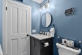 Blue bathroom with a white paneled door, round mirror above a dark vanity, and a bold light fixture; sink area has soap dispenser and towels nearby.