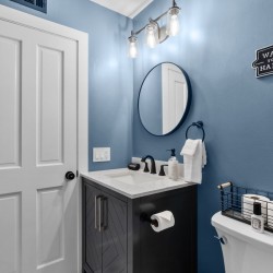 Blue bathroom with a white paneled door, round mirror above a dark vanity, and a bold light fixture; sink area has soap dispenser and towels nearby.