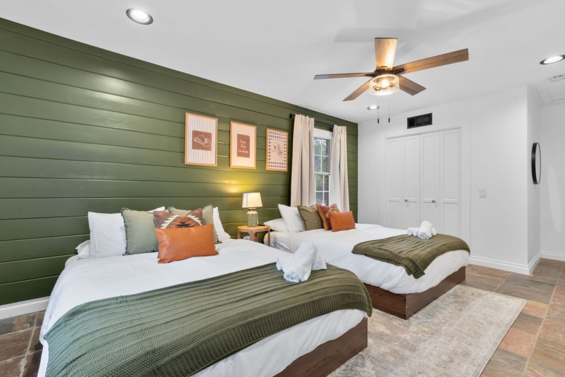 Bedroom with two twin beds, white linens, olive green shiplap wall, and a ceiling fan overhead.