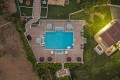 Aerial view of a rectangular blue pool with a tan tiled deck, surrounded by lounge chairs and umbrellas, with a building and green lawn nearby.