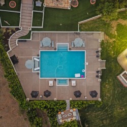 Aerial view of a rectangular blue pool with a tan tiled deck, surrounded by lounge chairs and umbrellas, with a building and green lawn nearby.