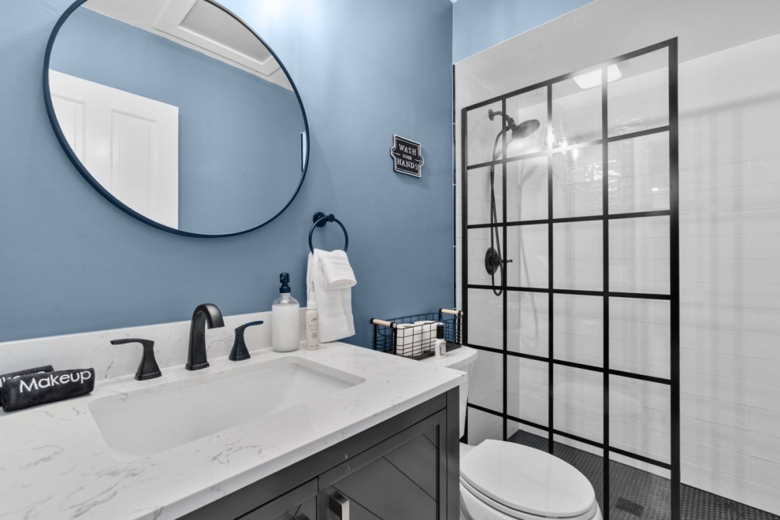 Blue-painted bathroom with a round wall mirror, white marble countertop, black faucet fixtures, towel ring, and a glass-walled shower with grid panels behind.