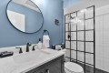 Blue-painted bathroom with a round wall mirror, white marble countertop, black faucet fixtures, towel ring, and a glass-walled shower with grid panels behind.