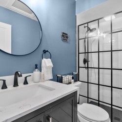 Blue-painted bathroom with a round wall mirror, white marble countertop, black faucet fixtures, towel ring, and a glass-walled shower with grid panels behind.