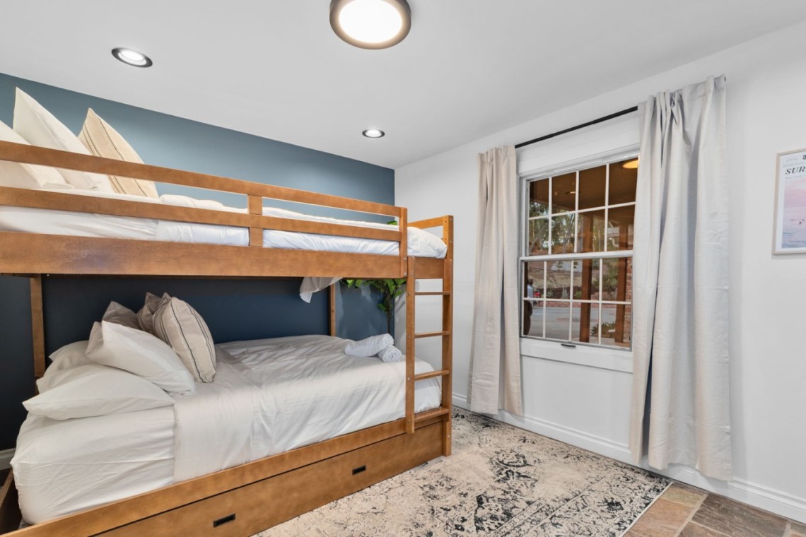 Wooden bunk bed with white linens in a small blue-accented bedroom, window with beige curtains to the right.