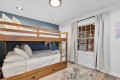 Wooden bunk bed with white linens in a small blue-accented bedroom, window with beige curtains to the right.