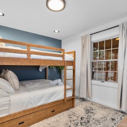 Wooden bunk bed with white linens in a small blue-accented bedroom, window with beige curtains to the right.
