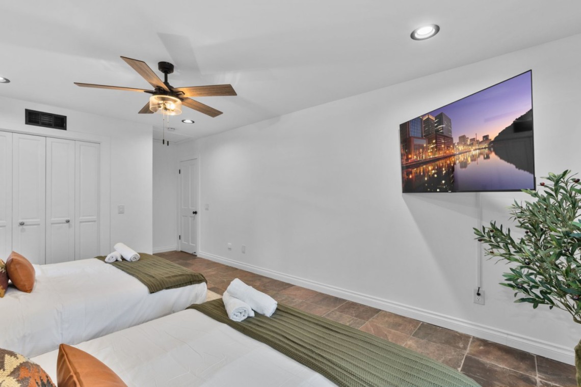 Twin bedroom with two single beds, white linens, olive-green throws, and orange pillows, plus a ceiling fan and wall-mounted TV on the right side.