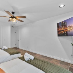 Twin bedroom with two single beds, white linens, olive-green throws, and orange pillows, plus a ceiling fan and wall-mounted TV on the right side.