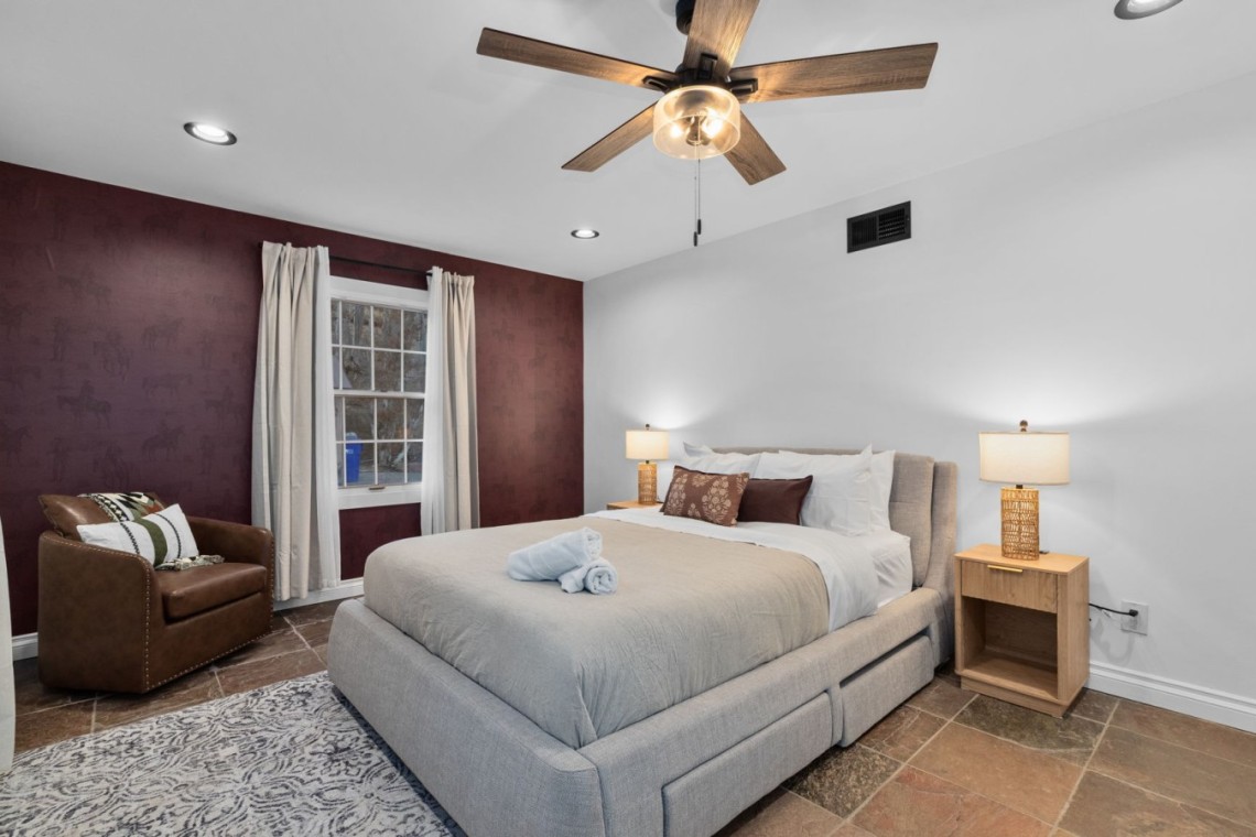 Modern bedroom with a gray upholstered bed, burgundy accent wall, window with light curtains, and two nightstands with lamps on either side of the bed.