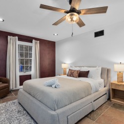 Modern bedroom with a gray upholstered bed, burgundy accent wall, window with light curtains, and two nightstands with lamps on either side of the bed.