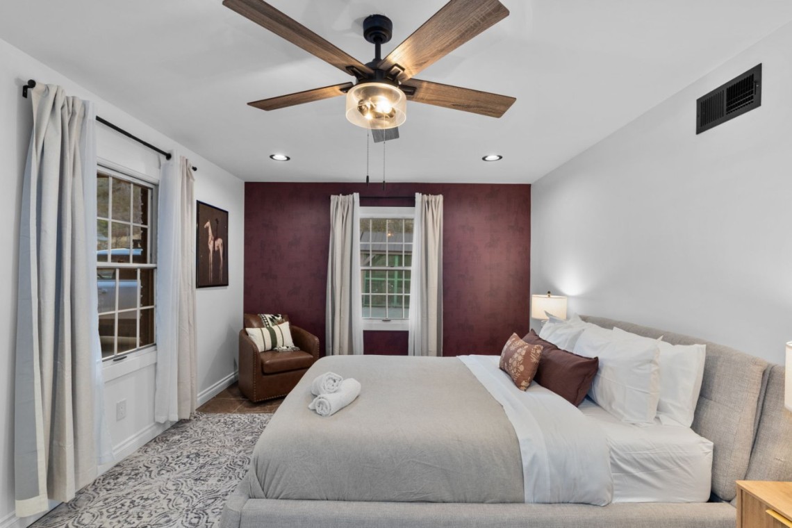 Bedroom with gray upholstered bed, white linens, and burgundy accent wall under a wooden ceiling fan.