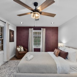 Bedroom with gray upholstered bed, white linens, and burgundy accent wall under a wooden ceiling fan.