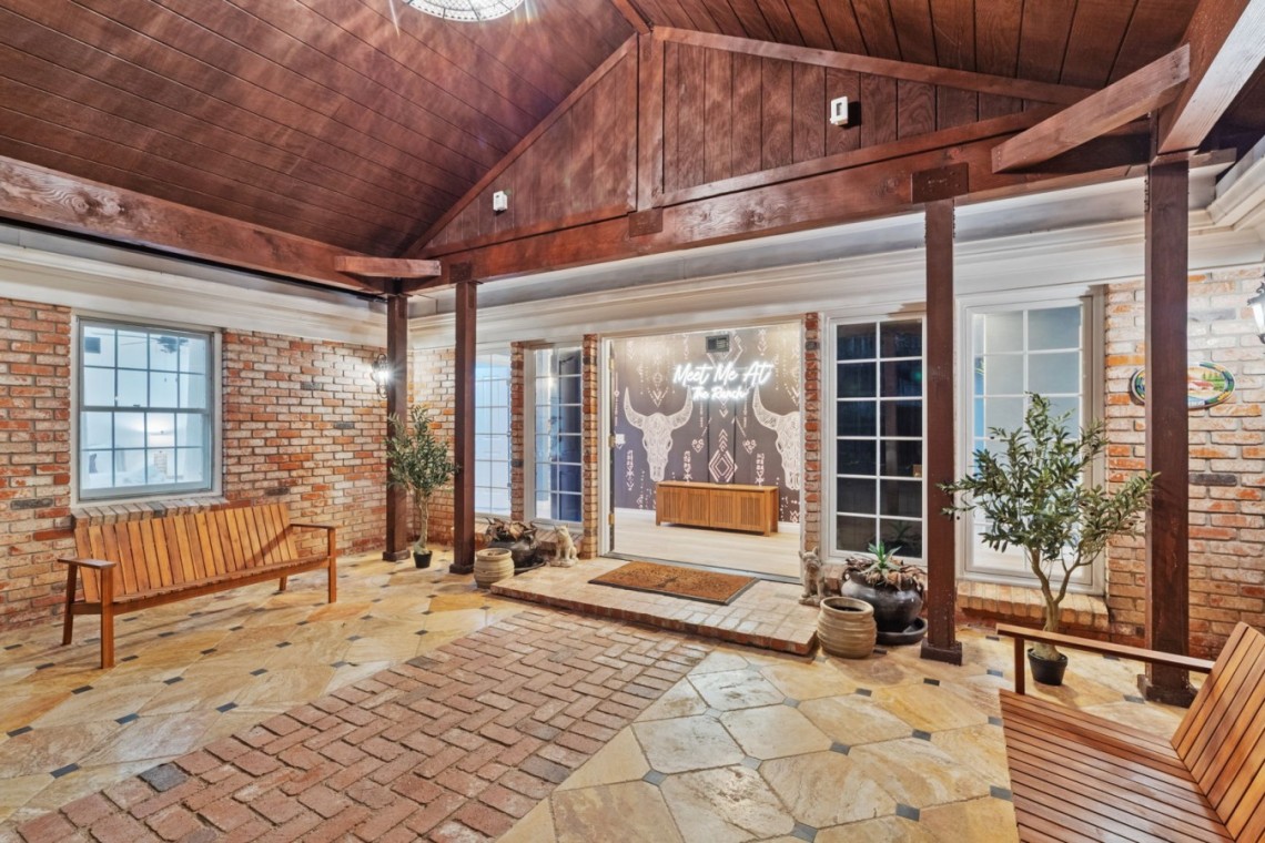 Brick-walled courtyard with a wood ceiling and timber beams, two wooden benches, potted plants, and glass doors opening to a mural-filled room.