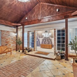 Brick-walled courtyard with a wood ceiling and timber beams, two wooden benches, potted plants, and glass doors opening to a mural-filled room.