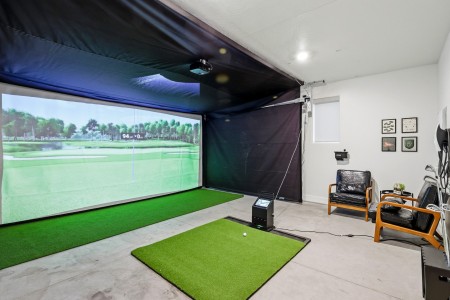 Indoor golf simulator setup: large screen showing a golf course, with green practice mats and a seating area nearby.