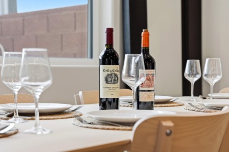 Table set for dinner with two wine bottles as centerpieces, empty wine glasses, plates, and utensils on a light tablecloth near a window.