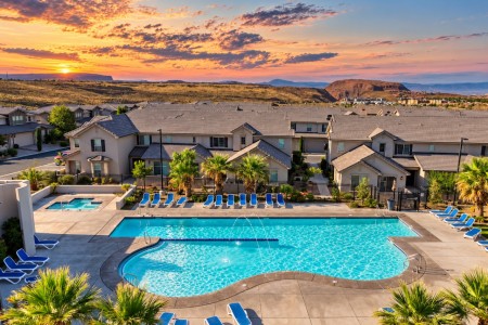 Aerial view of a resort-style apartment pool with blue loungers, palm trees, and beige townhomes at sunset.
