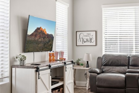 Living room with a wall-mounted flat-screen TV above a white rustic cabinet, snacks and drinks on the surface, and a gray leather sofa nearby, plus a framed “Make yourself at home” sign.