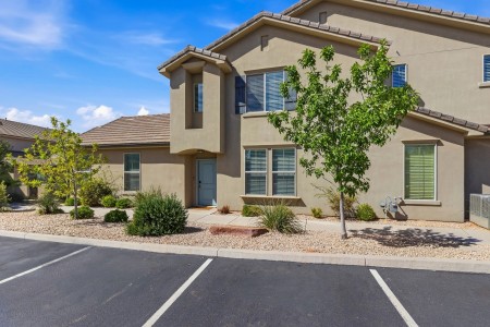 Two-story beige stucco townhouse with a blue-gray front door, arched entry, and large front windows, surrounded by desert landscaping and small trees; a paved parking lot is in the foreground.
