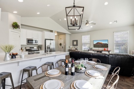Open-concept kitchen and dining area with a wooden table set for six, wine bottles as a centerpiece, white cabinets, and stainless-steel appliances nearby.